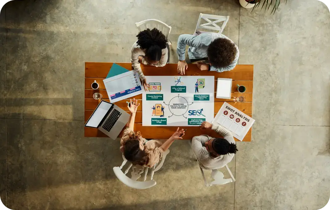 Four individuals are seated around a wooden table, engaged in a discussion. The table contains a laptop, documents, and a diagram related to marketing and SEO strategies.